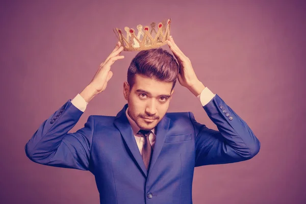 紫色の背景に金色の冠を頭の上保持青いスーツを着た若い魅力的な男 — ストック写真