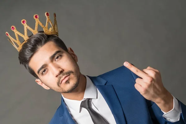 紺のスーツと灰色の背景に金の王冠を示す中指で若い魅力的な男 — ストック写真