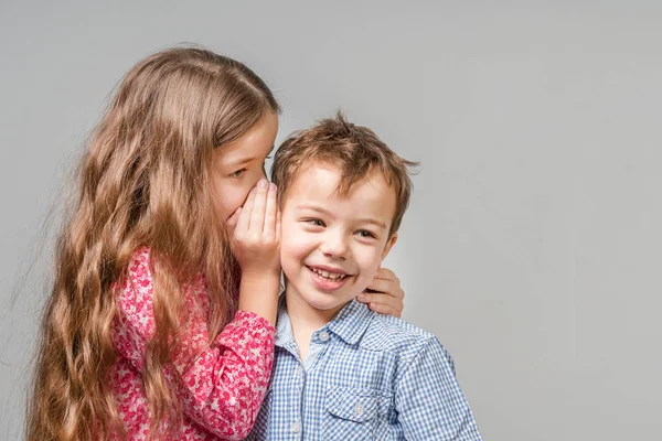 Girl in a red dress whispers in the ear a boy in a blue shirt on a gray background. Isolated