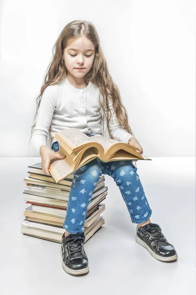 Cute girl with a book sitting on a pile of books on a white background