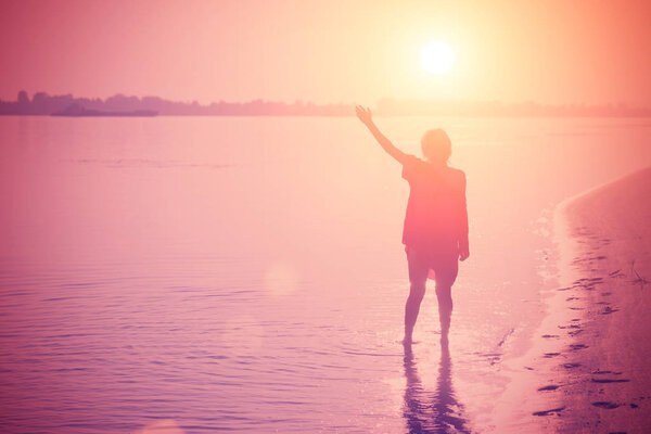 Girl teenager standing in the water with a raised hand at pink sunset