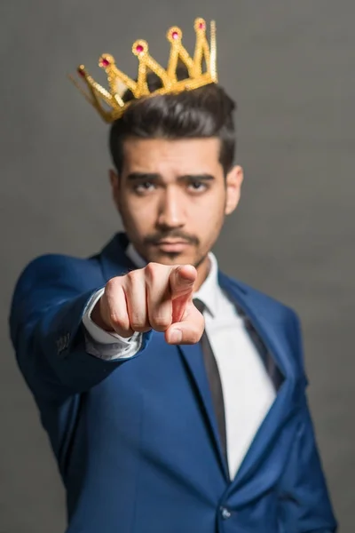 Young attractive man in a blue suit with a crown on his head shows a finger forward on a gray background