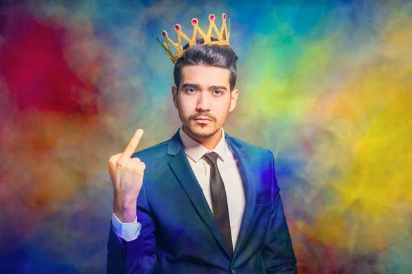 Young attractive man in a blue suit with a crown on his head showing middle finger on a colorful background