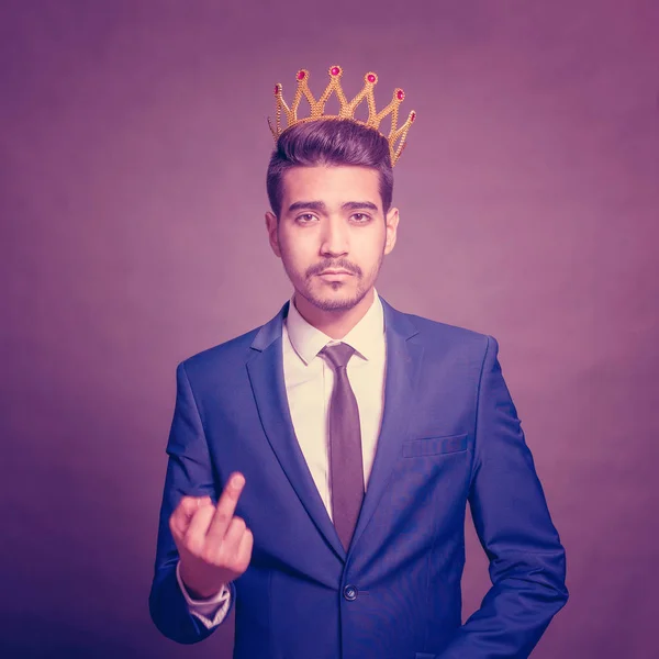 Young attractive man in a blue suit with a crown on his head showing middle finger on a purple background