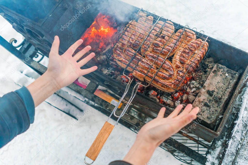 Man warms his hands over the brazier with sausages grilled in winter