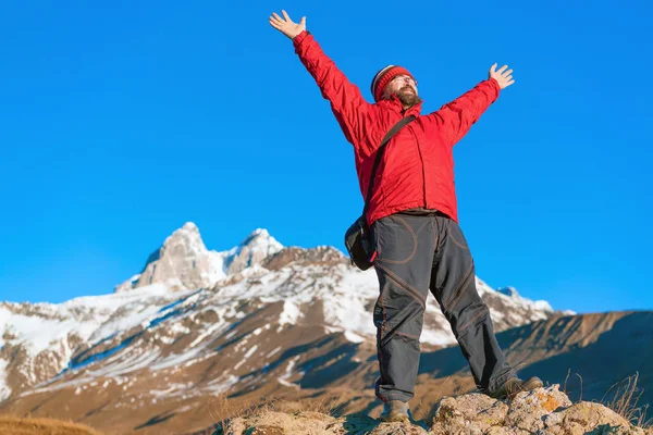 Vousatý muž v červeném kabátku s rukama nahoře v horách. — Stock fotografie