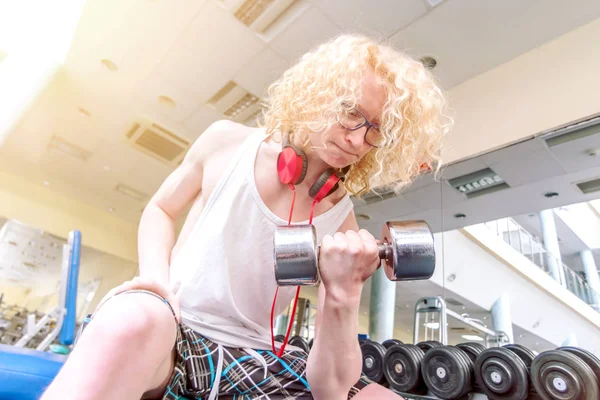 Gym. Curly blond guy in a white vest lifting dumbbell with a str