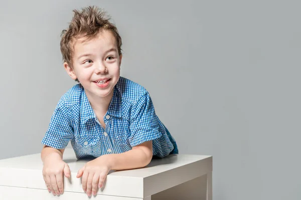 Rozkošný usměvavý chlapec u stolu na šedém pozadí studia — Stock fotografie