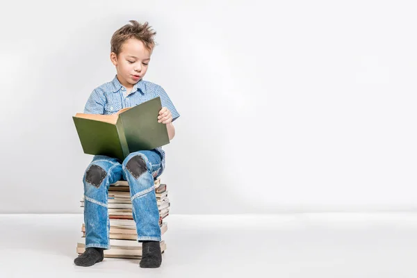 Lindo chico sentado en una pila de libros y leyendo un libro . — Foto de Stock