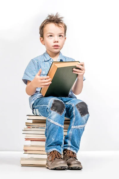 Lindo chico sentado en un montón de libros y sosteniendo un libro . — Foto de Stock