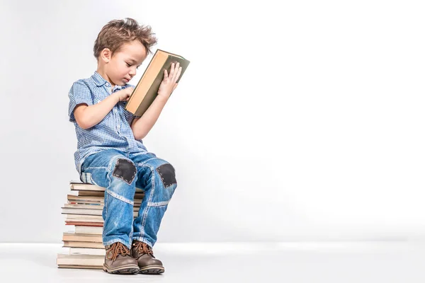 Un chico lindo está sentado en una pila de libros y está considerando una b — Foto de Stock
