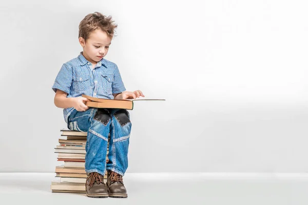 Lindo chico se sienta en una pila de libros y lee un libro sobre una ba blanca — Foto de Stock