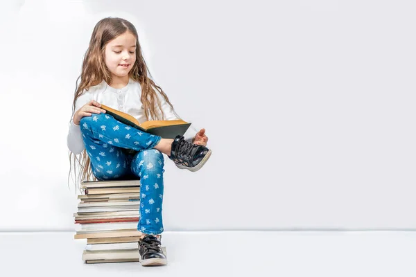 Una chica bonita está sentada en una pila de libros y leyendo un libro — Foto de Stock