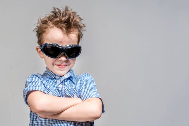 Gri arka plan üzerinde katlanmış kolları ile güneş gözlüğü gülümseyen çocuk.
