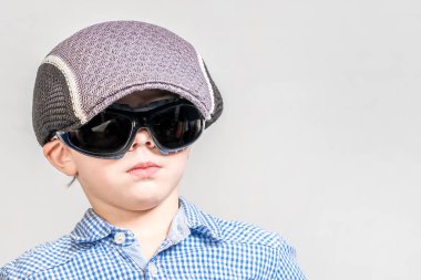 Gri bir arka plan üzerinde şapkalı ve güneş gözlüğü takan ciddi bir çocuk. ısola