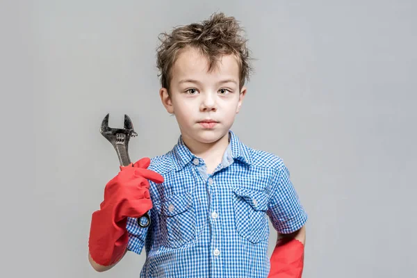 Cute boy holding a wrench in red gloves on a gray background. Is