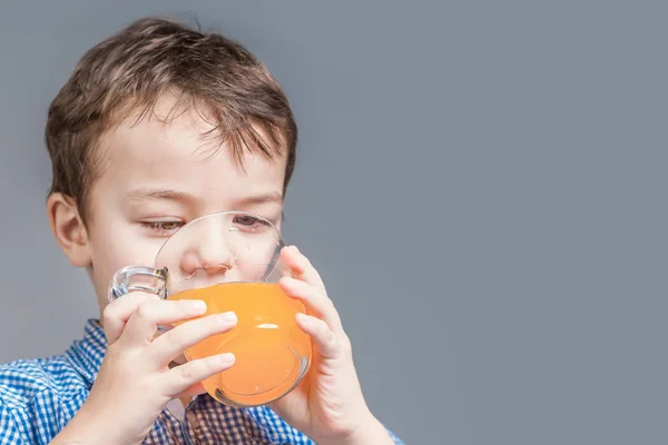 Mavi gömlekli çocuk gri bir arka planda portakal suyu içiyor. — Stok fotoğraf