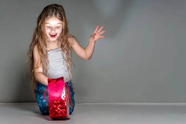 Sorprendida chica feliz mira en una bolsa de regalo roja en un fondo gris —  Fotos de Stock