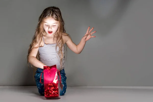 Chica sorprendida mira en una bolsa de regalo brillante rojo en un backgro gris —  Fotos de Stock