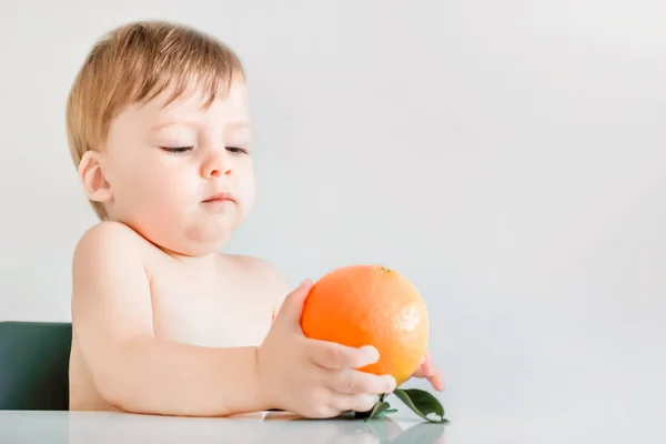 Sevimli sarışın bebek masada otururken bir portakal tutar. — Stok fotoğraf