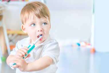 Cute blue-eyed blond baby brushing his teeth with a toothbrush. clipart