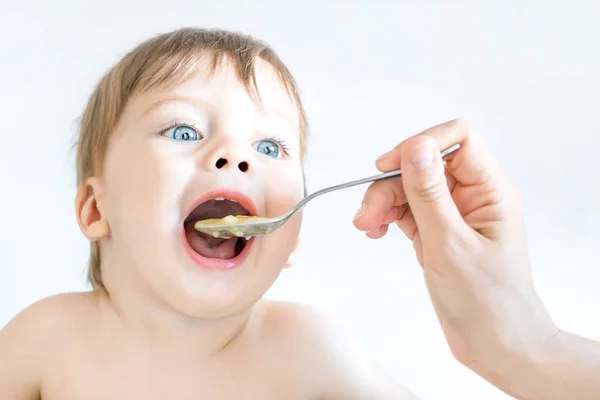 Lindo bebé de ojos azules está comiendo con una cuchara, con la ayuda de mamá . — Foto de Stock