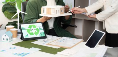 Bir ESG projesinde işbirliği yapan profesyoneller, modern bir çalışma alanında bir model bina ve dijital cihazlar sergiliyorlar.
