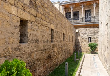 Bakü, Azerbaycan, 07.25.2019. Antik, ortaçağ, açıkları olan kale duvarı ve gece aydınlatması için lambaları olan yeşil bir çimen. Duvara yakın bir apartmanın verandası.