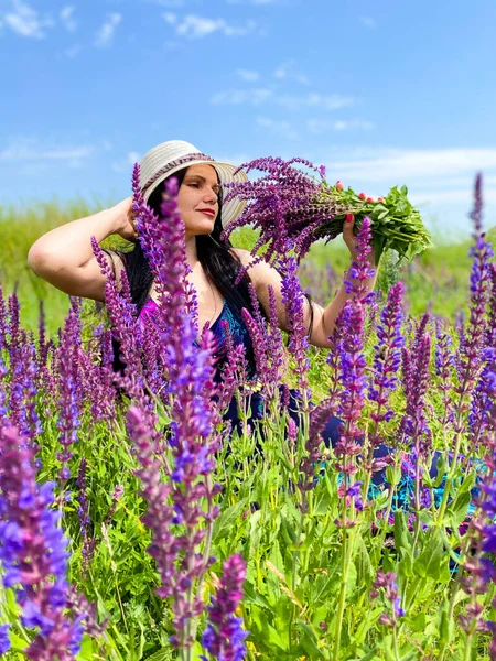 A fair-skinned woman with dark long hair in a straw hat, in a field among purple flowers. Free space.