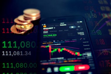 Bir akıllı telefon, arka planda dalgalanan pazar grafikleri ile birlikte altın sikkelerin yanında şifreli para ticareti bilgisini görüntüler.