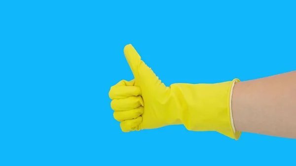 Primo piano della mano femminile in guanto di gomma di lattice protettivo giallo che mostra i pollici verso l'alto segno, gesto contro lo sfondo blu, fatto concetto di lavori domestici — Foto Stock