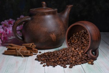 Çamur çaydanlığının arka planında kahve çekirdekleri, şeker küpleri, tarçın çubukları ve ahşap masada kiraz çiçekleri olan ters çevrilmiş kupa, kahve hala hayat dolu.