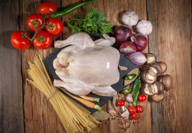Tavuk pişirmek için taze ürünler. Tavuk, havuç, mantar, soğan, körfez yaprağı, domates, biber, dereotu, maydanoz, makarna, dökme demir kap, ahşap masa, yakın çekim, üst manzara, kırsal tarz.