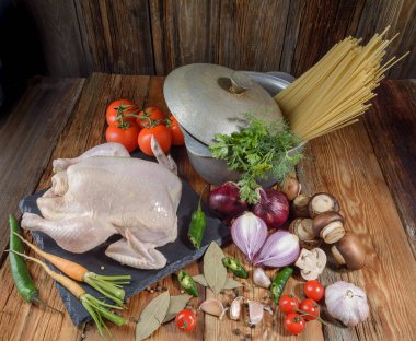 Tavuk pişirmek için taze ürünler. Tavuk, havuç, mantar, soğan, körfez yaprağı, domates, biber, dereotu, maydanoz, makarna, dökme demir kap, ahşap masa, yakın çekim, üst manzara, kırsal tarz.