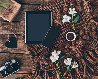 Düz ahşap masanın üzerinde atış örme eşarp, çiçekler, fincan kahve, hediye kutusu, fotoğraf makinesi, akıllı telefon ve dijital tablet yatıyordu