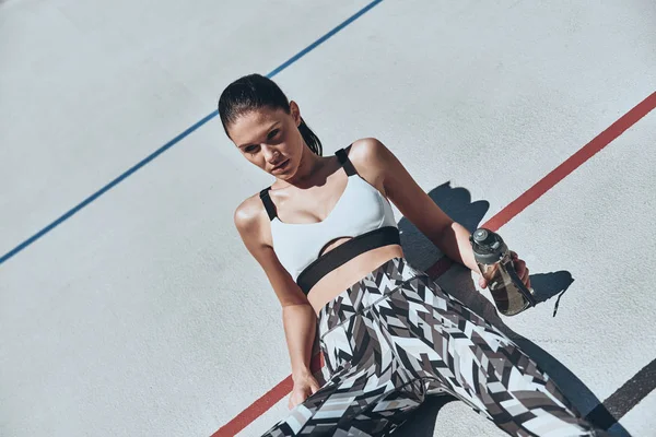 brunette woman in sports clothing resting on running track stadium and holding plastic bottle with water