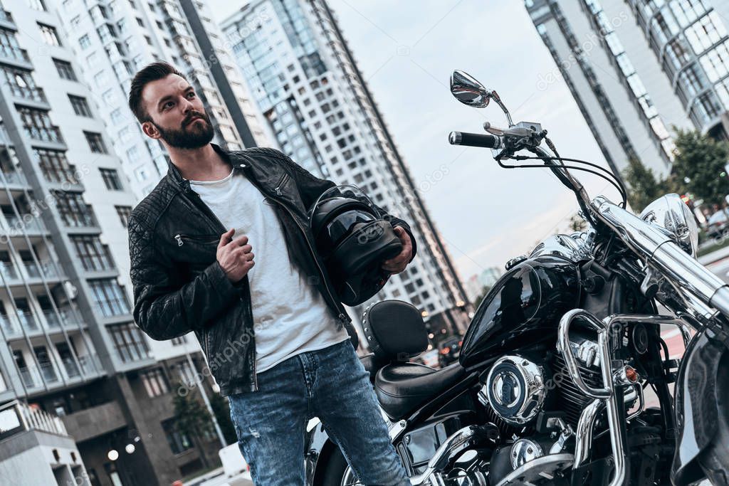 handsome rockabilly motorcyclist man in leather jacket standing near motorbike outdoors