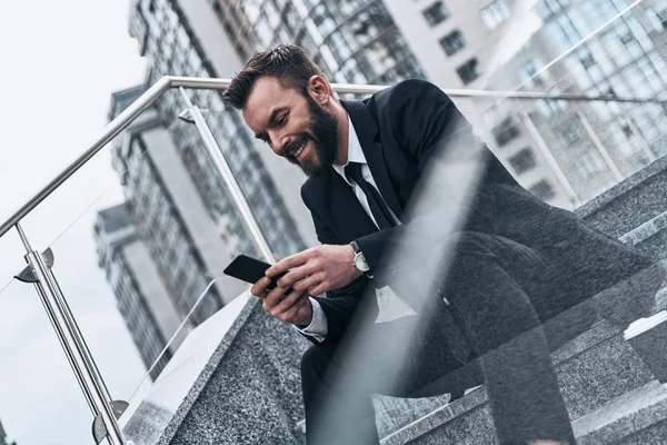 smiling man in suit using smart phone while sitting on stairs outdoors