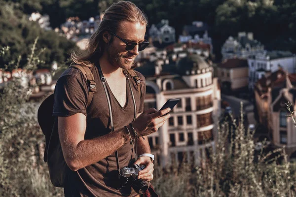 stylish man with camera using smart phone while standing on hill outdoors