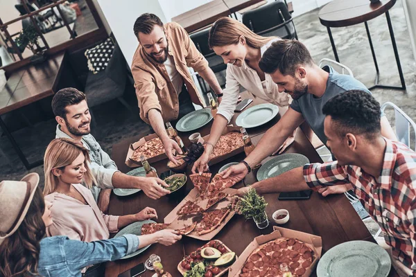 friends in casual clothing picking pizza and smiling while having dinner party