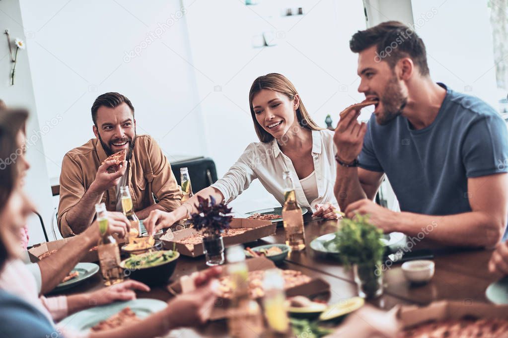 hungry young people in casual clothing eating and smiling while having a dinner party indoors