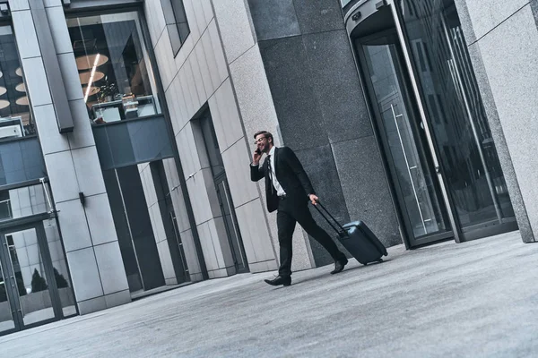 愉快的商人在西装拉扯行李和谈论移动电话 当步行在室外在办公室大厦 — 图库照片