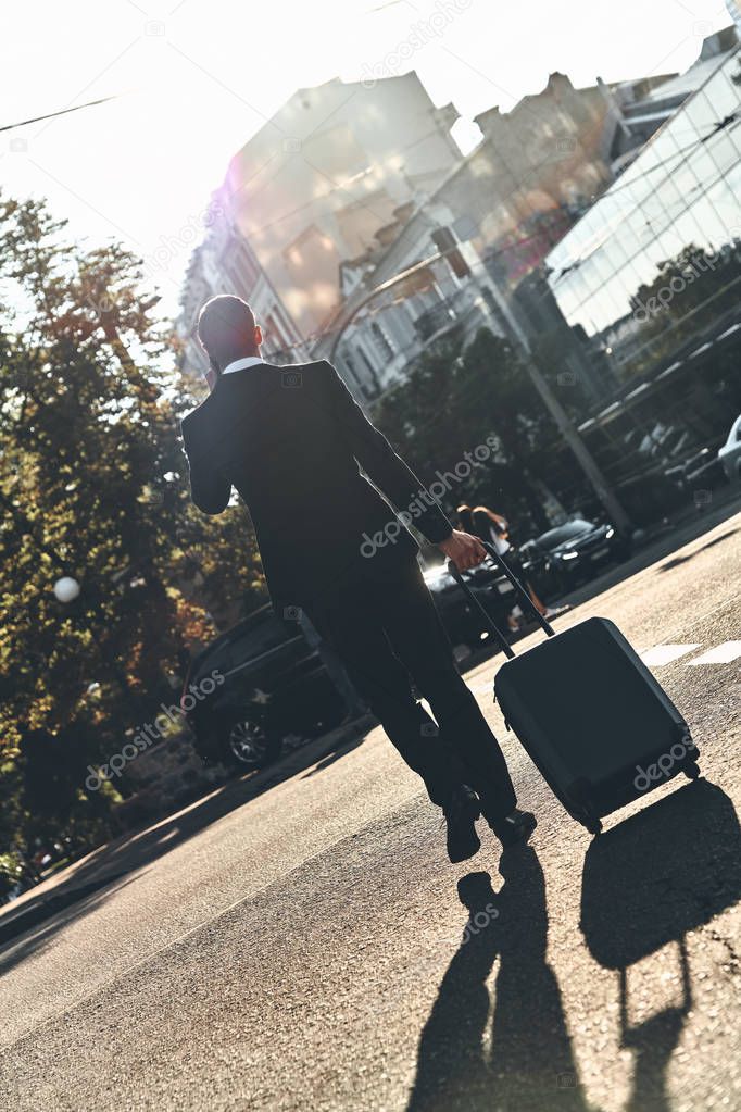 back view of businessman in suit walking in city street with travel luggage 