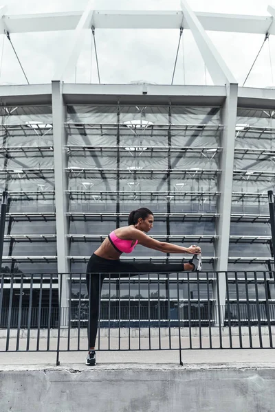 sportive woman in pink top stretching legs outdoors at railings of stadium