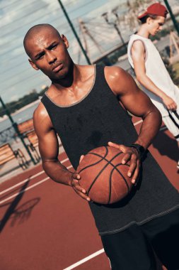 serious young African man in sports clothing looking at camera and holding basketball ball clipart