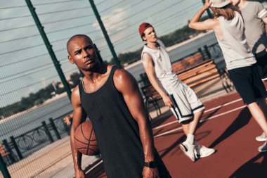sportive African man looking away while holding basketball ball on court with friends clipart