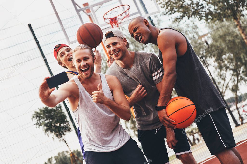 group of smiling men in sports clothing taking selfie on mobile phone while posing on basketball arena court