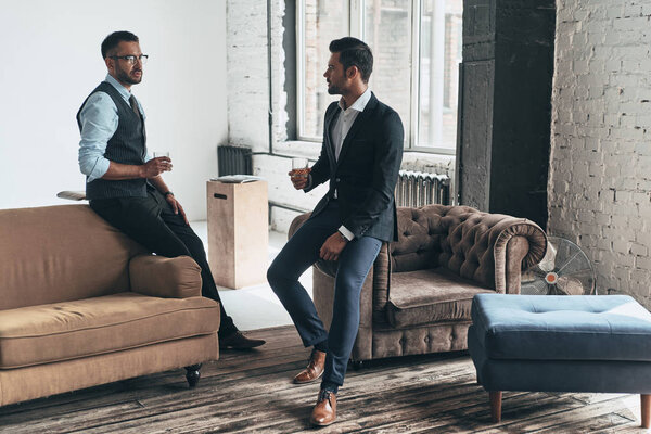 полная длина двух молодых красивых мужчин в костюмах, держащих очки и смотрящих друг на друга во время отдыха в помещении
