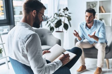 man having therapy session with psychologist, doctor writing notes in diary with pen      clipart