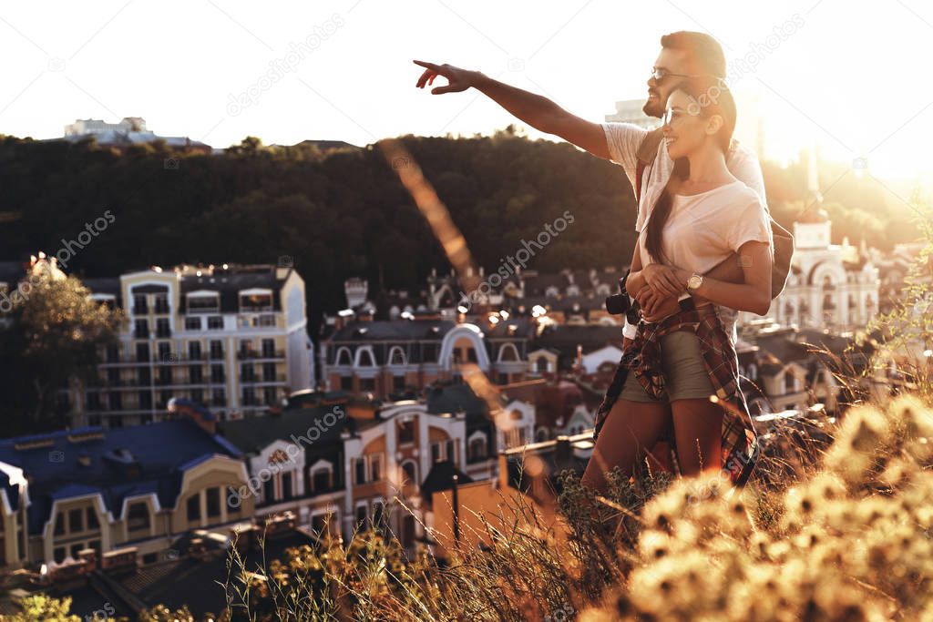 beautiful young couple embracing and smiling outdoors on hills, man pointing with finger 